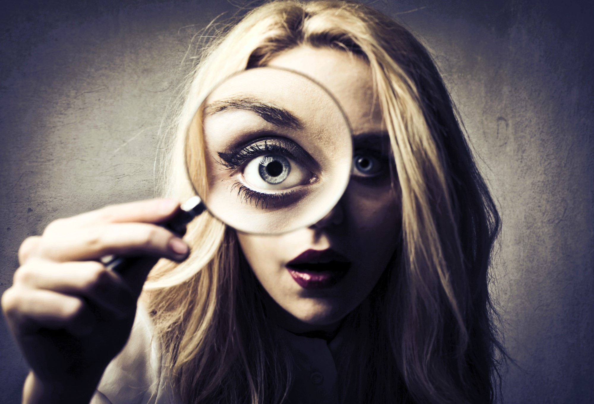 A woman with long blonde hair looks closely through a magnifying glass, which enlarges one of her eyes. The background is a plain, textured wall. Her expression is curious and slightly dramatic.