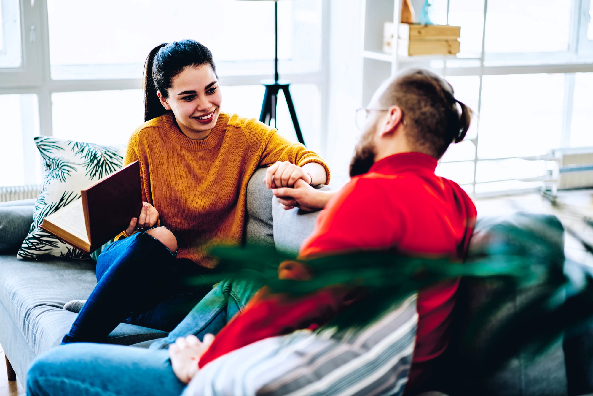 A woman in a yellow sweater is sitting on a couch, holding a book and smiling at a man in a red shirt with glasses. They are engaged in a conversation in a bright, cozy living room with pillows and a large window behind them.