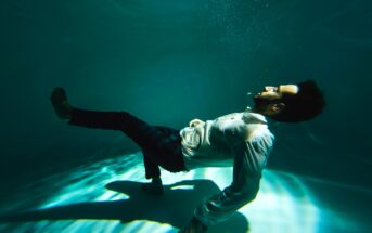 A man in a white shirt and dark pants floats underwater, partially reclining with knees bent and arms resting at his sides. Sunlight filters through the water, creating patterns on the pool floor. Bubbles rise from his mouth as he looks upward.