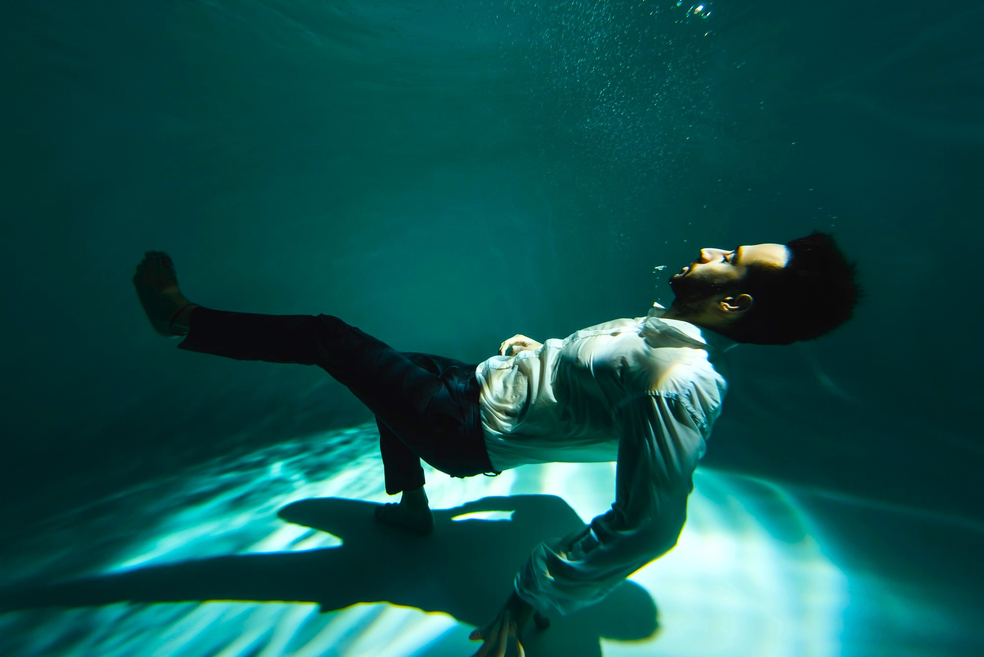 A man in a white shirt and dark pants floats underwater, partially reclining with knees bent and arms resting at his sides. Sunlight filters through the water, creating patterns on the pool floor. Bubbles rise from his mouth as he looks upward.