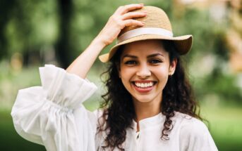 A woman with curly hair smiles brightly, wearing a straw hat and a white blouse. She is outdoors with a blurred green background. Her hand is gently touching the hat, and she appears joyful and relaxed.