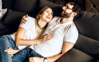 A couple is relaxing on a couch, both wearing white shirts and jeans. The woman is lying against the man's chest, looking up and smiling, while he looks down at her with a gentle expression. They appear comfortable and happy together.