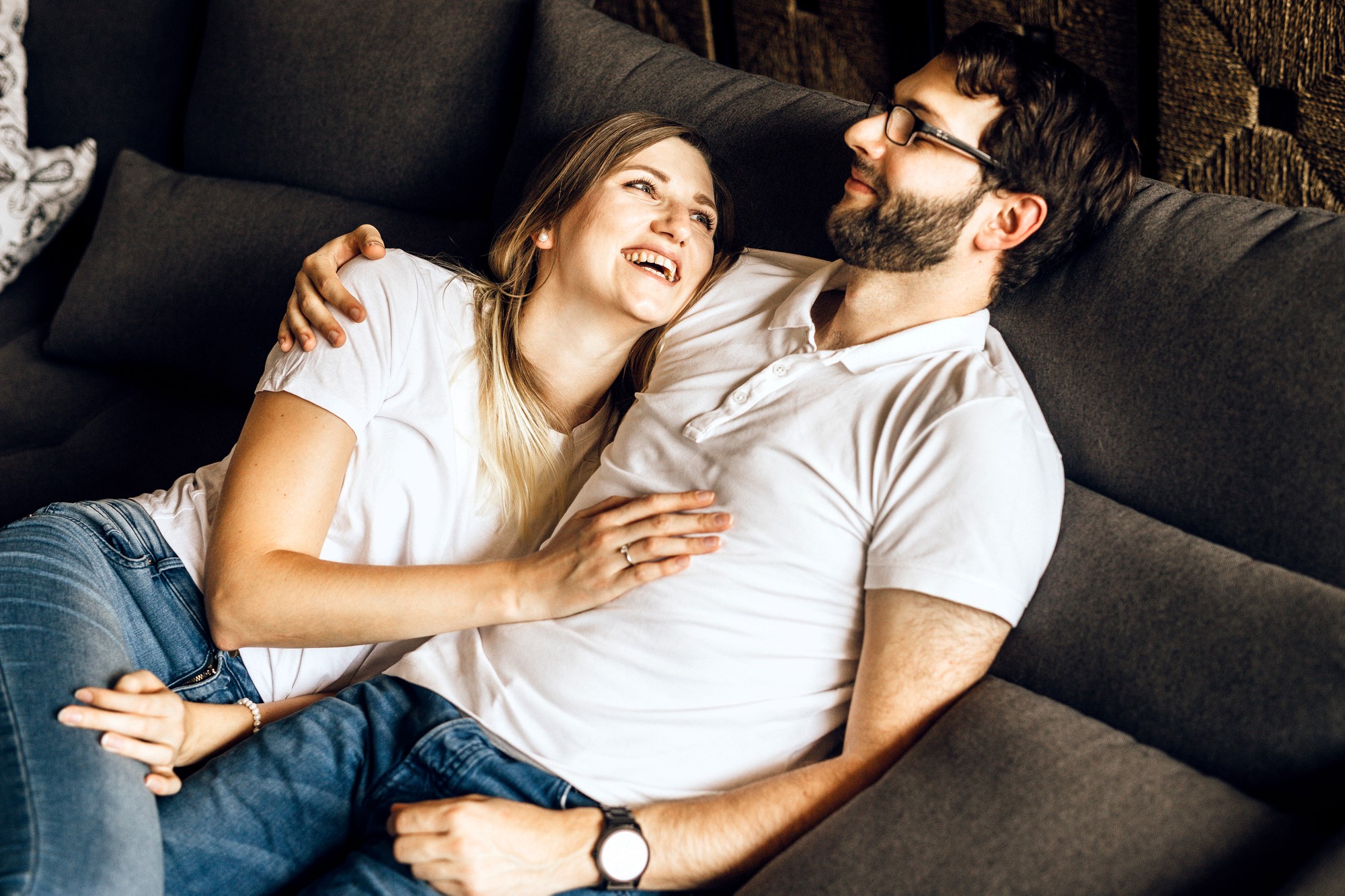 A couple is relaxing on a couch, both wearing white shirts and jeans. The woman is lying against the man's chest, looking up and smiling, while he looks down at her with a gentle expression. They appear comfortable and happy together.