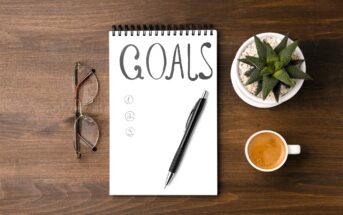 A spiral notebook with "GOALS" written at the top, list numbered 1 to 3, and a pen rests on it. Nearby are eyeglasses, a small potted succulent, and a cup of espresso, all on a wooden surface.