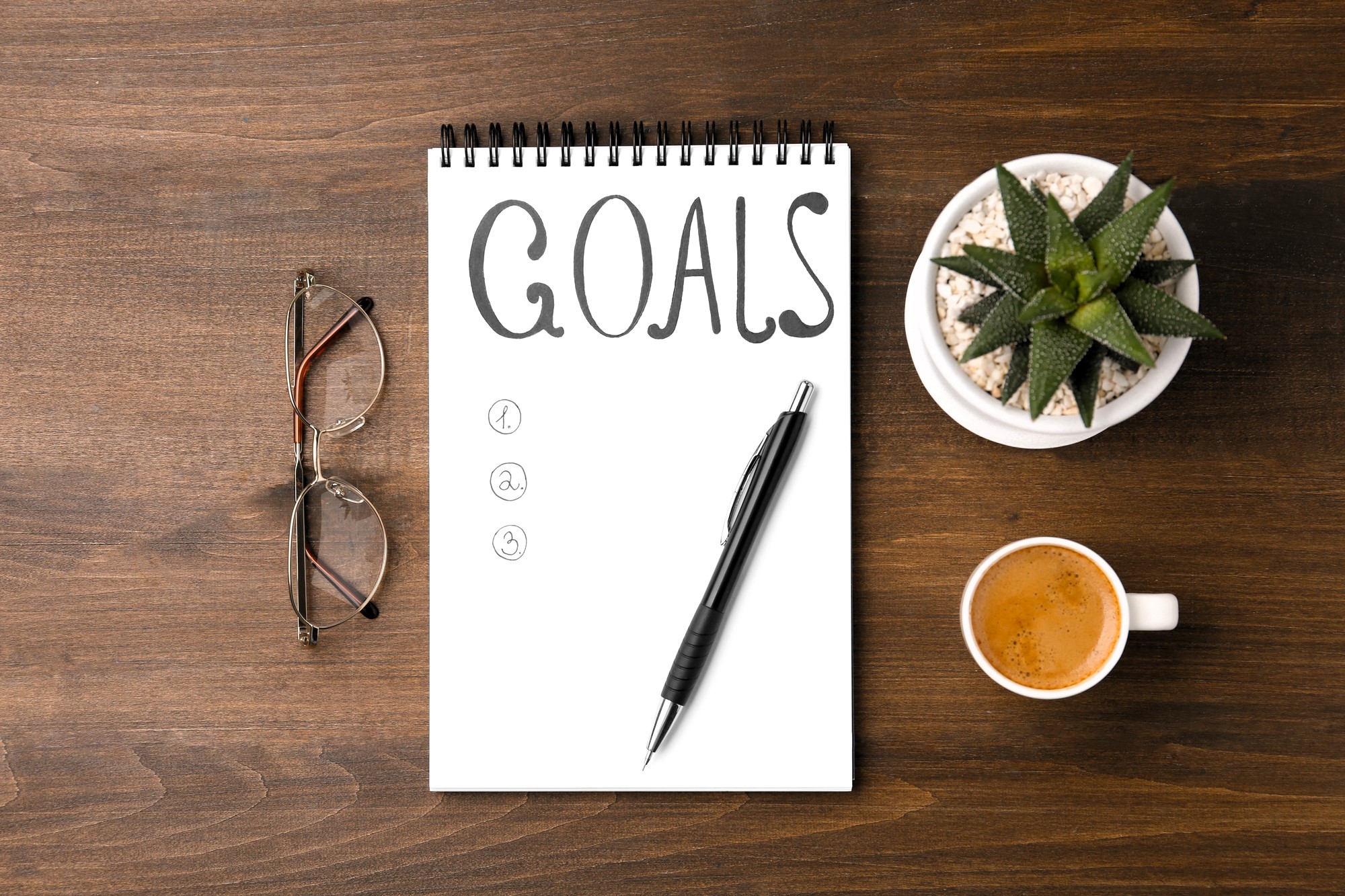 A spiral notebook with "GOALS" written at the top, list numbered 1 to 3, and a pen rests on it. Nearby are eyeglasses, a small potted succulent, and a cup of espresso, all on a wooden surface.