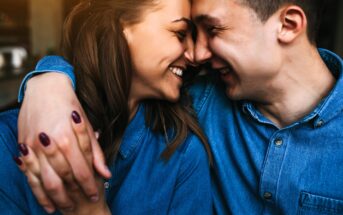 A couple wearing denim shirts smiles and leans their foreheads together. They have their arms around each other, showcasing a close and affectionate moment. The background is softly blurred, emphasizing their joyful expressions.