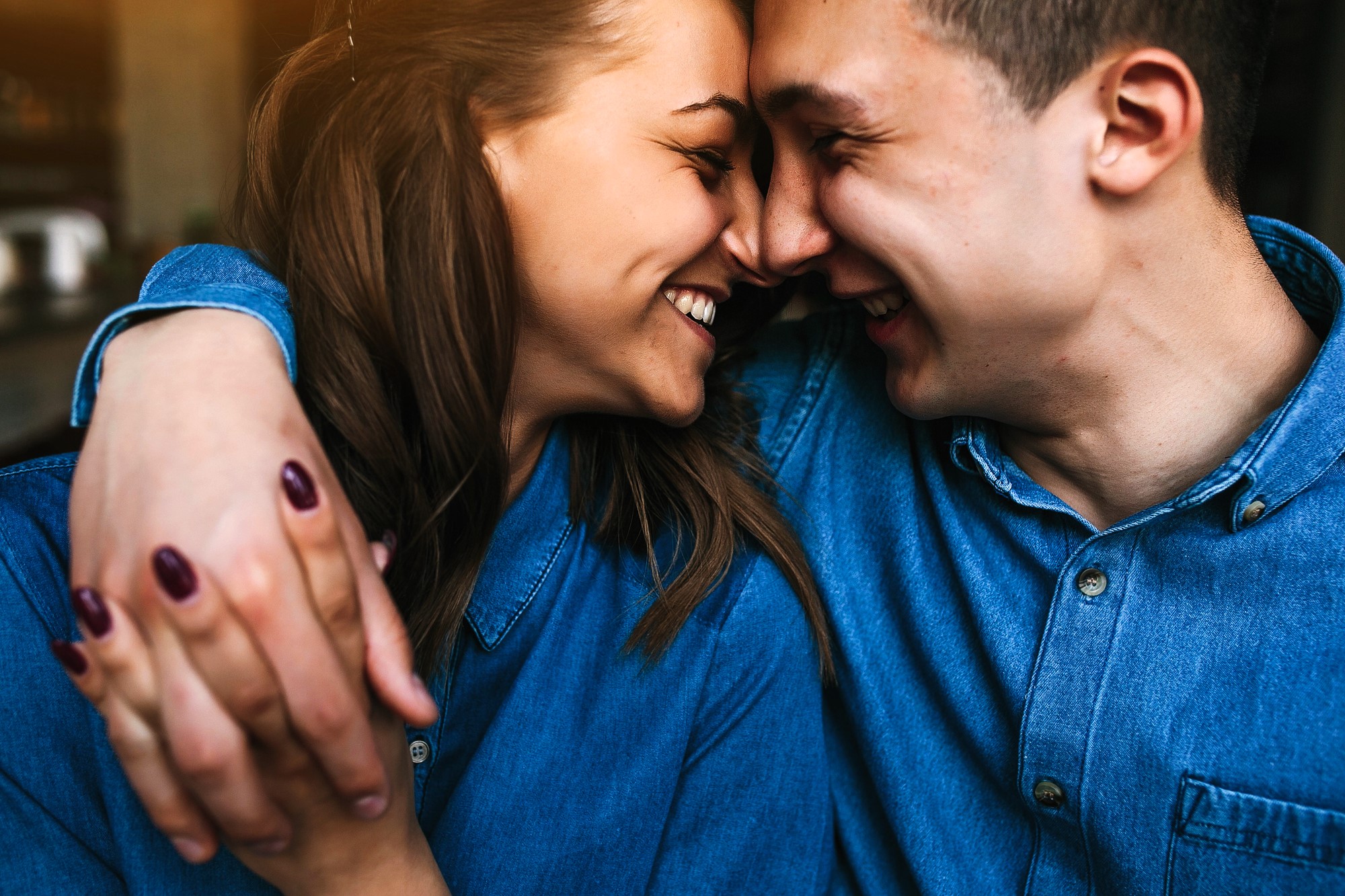 A couple wearing denim shirts smiles and leans their foreheads together. They have their arms around each other, showcasing a close and affectionate moment. The background is softly blurred, emphasizing their joyful expressions.