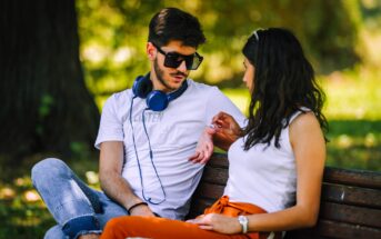 A man wearing sunglasses and headphones sits on a bench with a woman in a park. They are talking, and the woman gestures while making eye contact. Both wear casual clothes, and the background shows blurred greenery.