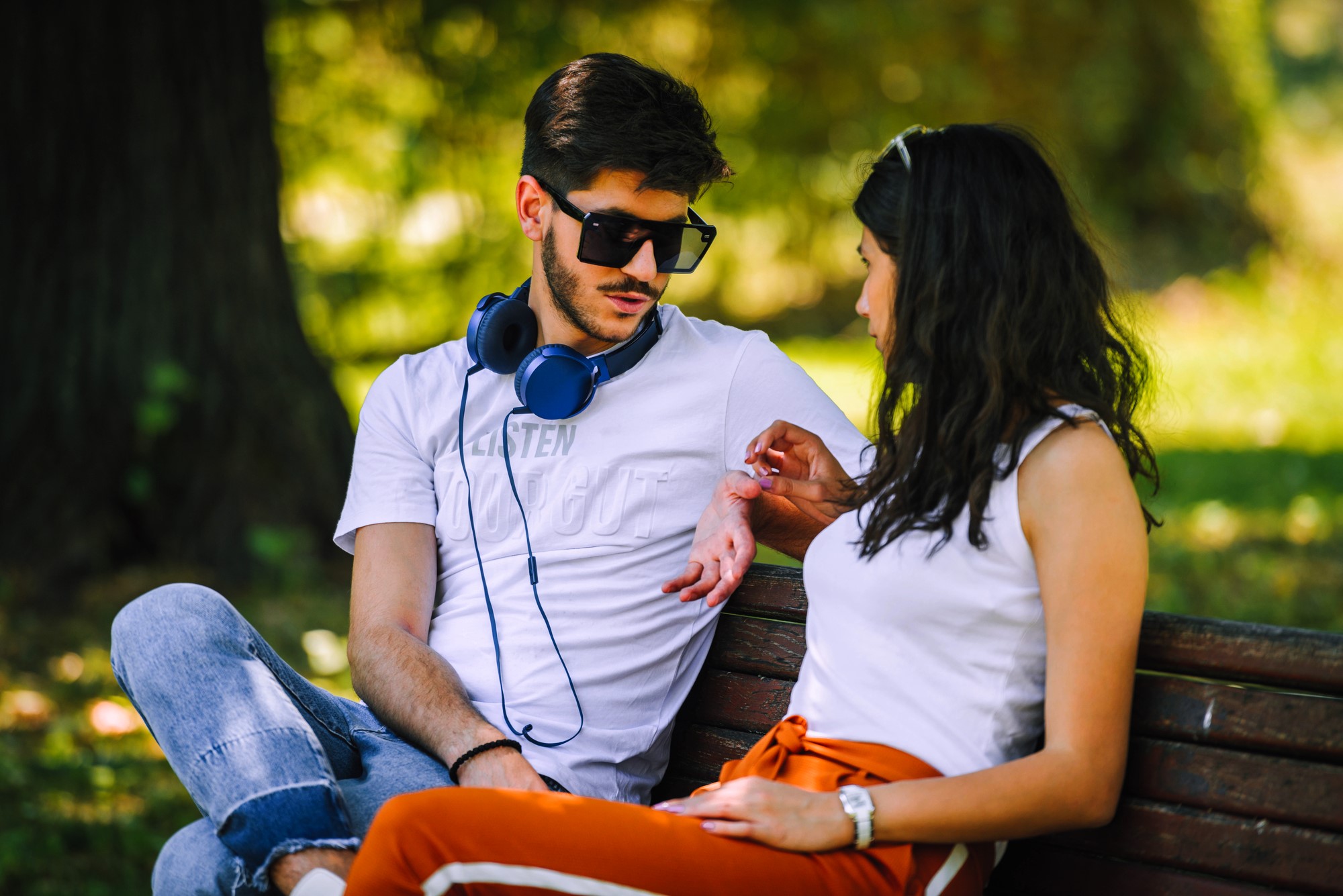 A man wearing sunglasses and headphones sits on a bench with a woman in a park. They are talking, and the woman gestures while making eye contact. Both wear casual clothes, and the background shows blurred greenery.