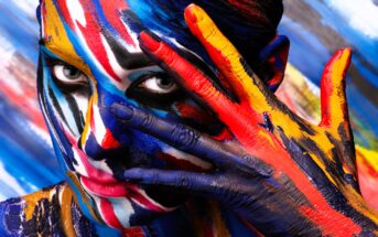 A person with vibrant multicolored paint on their face and fingers, including blue, red, yellow, and white, peeks through their open hand against a similarly painted background. The image radiates creativity and expression.