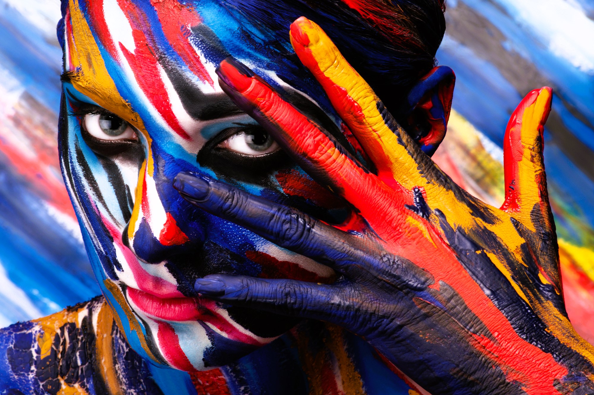 A person with vibrant multicolored paint on their face and fingers, including blue, red, yellow, and white, peeks through their open hand against a similarly painted background. The image radiates creativity and expression.