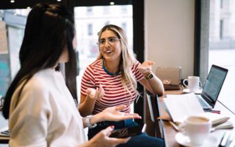 Two women are sitting at a cafe table, talking and smiling. One has long blonde hair and wears glasses with a red striped shirt. The other has dark hair and holds a phone. A laptop, notebook, and coffee cups are on the table.