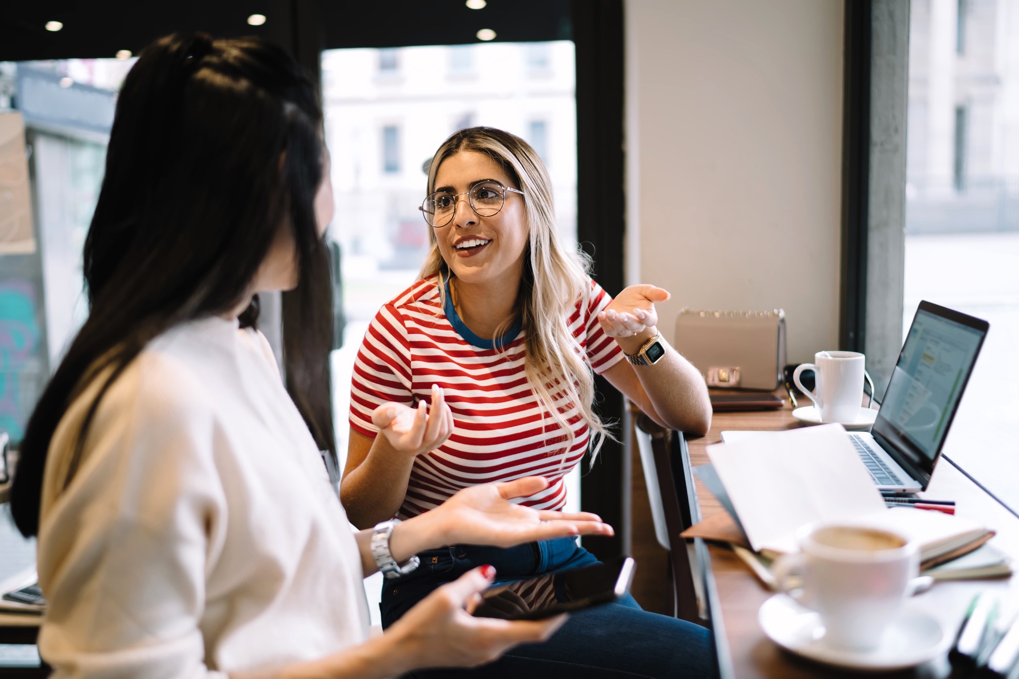 Two women are sitting at a cafe table, talking and smiling. One has long blonde hair and wears glasses with a red striped shirt. The other has dark hair and holds a phone. A laptop, notebook, and coffee cups are on the table.