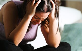 A woman sitting on a couch clutches her head with both hands, appearing stressed or upset. She is wearing a sleeveless top and dark pants, with a blurred cushion visible in the background.