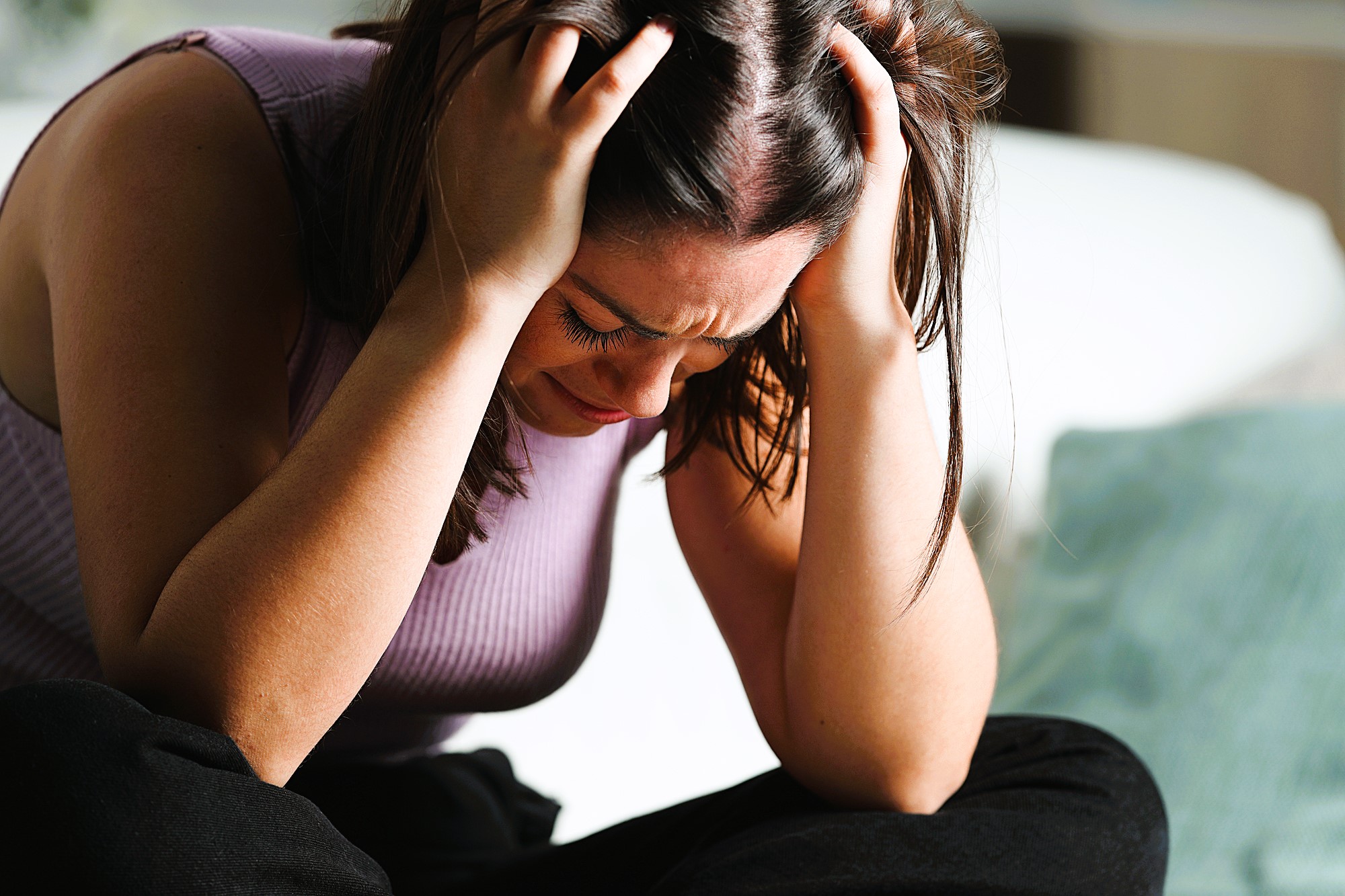 A woman sitting on a couch clutches her head with both hands, appearing stressed or upset. She is wearing a sleeveless top and dark pants, with a blurred cushion visible in the background.