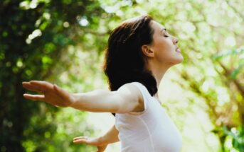 A woman with dark hair, wearing a white shirt, stands outdoors with her arms outstretched and eyes closed. She appears to be enjoying the fresh air. The background is blurred and filled with sunlight filtering through green leaves.