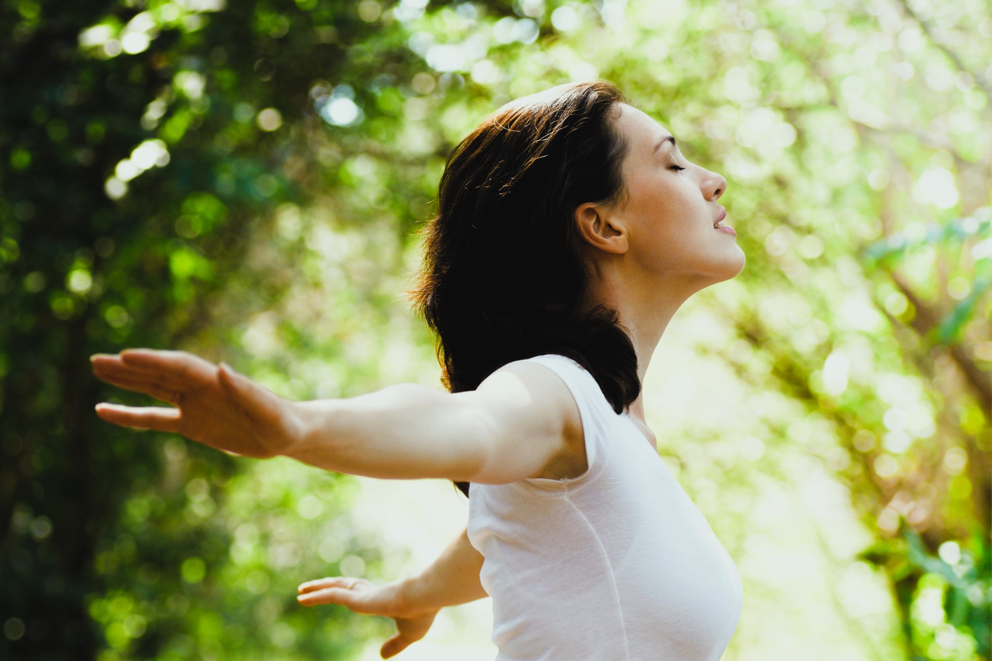 A woman with dark hair, wearing a white shirt, stands outdoors with her arms outstretched and eyes closed. She appears to be enjoying the fresh air. The background is blurred and filled with sunlight filtering through green leaves.