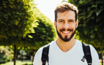 A smiling man with a beard and white t-shirt stands outdoors in front of lush green trees. He is wearing a backpack and the sun is shining brightly.