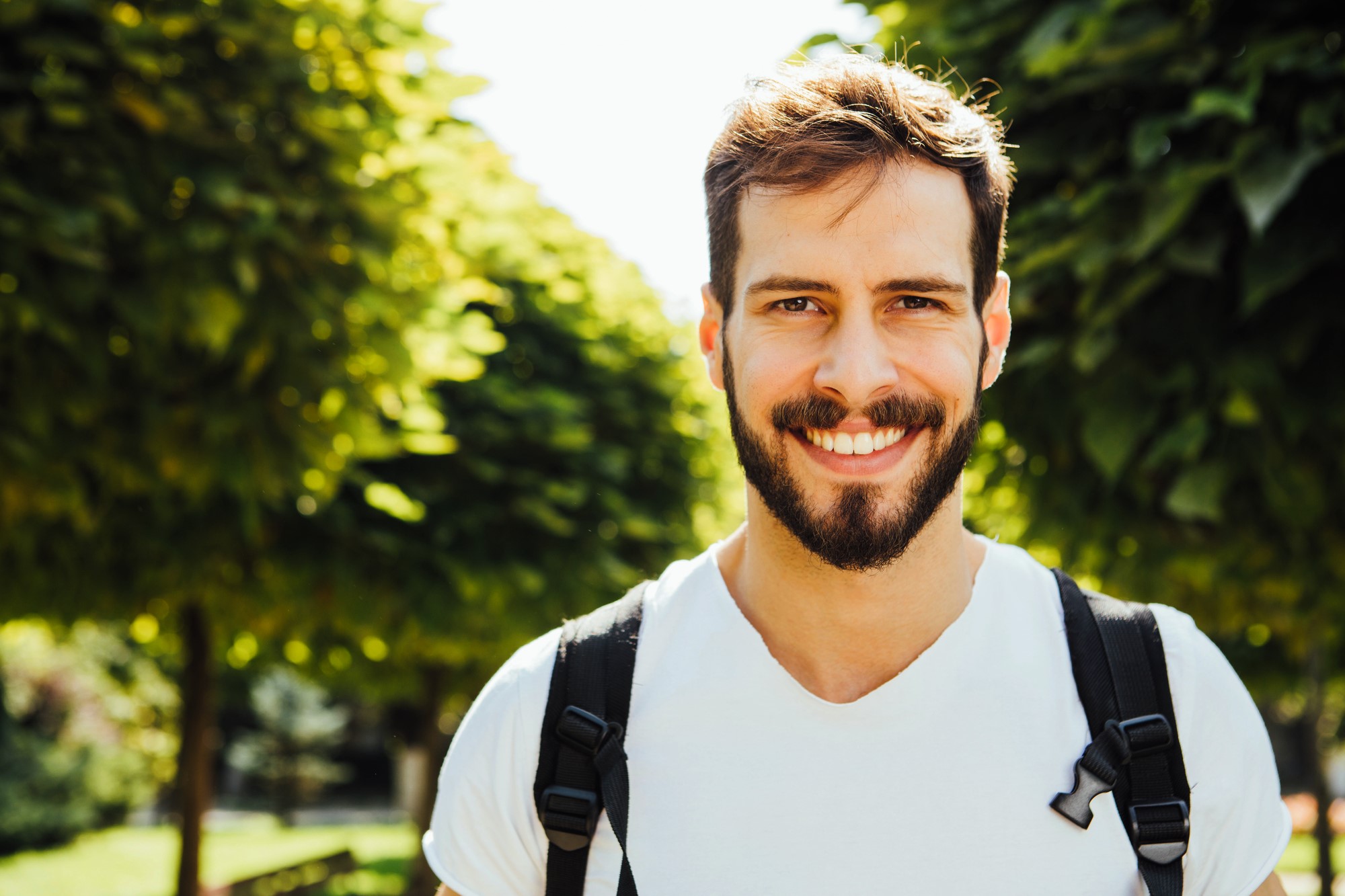 A smiling man with a beard and white t-shirt stands outdoors in front of lush green trees. He is wearing a backpack and the sun is shining brightly.