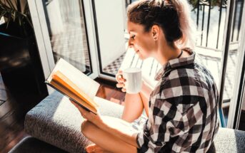 A person sits on a couch in sunlight, reading a book and holding a white mug. They wear a plaid shirt and have light-colored hair tied back. Large windows and a balcony are in the background.