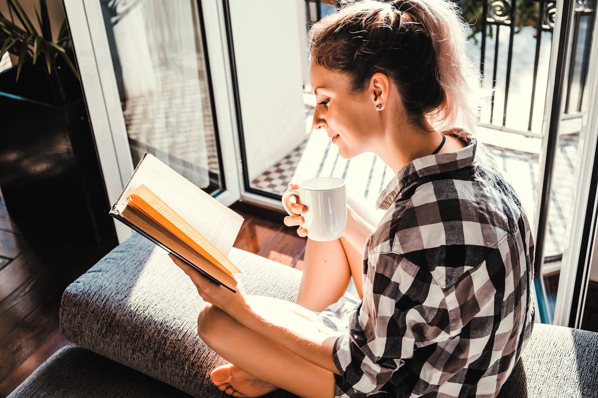 A person sits on a couch in sunlight, reading a book and holding a white mug. They wear a plaid shirt and have light-colored hair tied back. Large windows and a balcony are in the background.