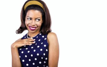 A person with dark hair and a yellow headband is smiling mischievously and looking to the side. They are wearing a navy blue dress with white polka dots and a pearl necklace, with one hand raised to their chest. The background is white.