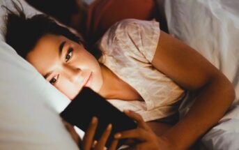 A woman is lying in bed at night, holding and looking at a smartphone. Her face is softly illuminated by the screen. She appears relaxed, with her head resting on a pillow. Another person is lying beside her.