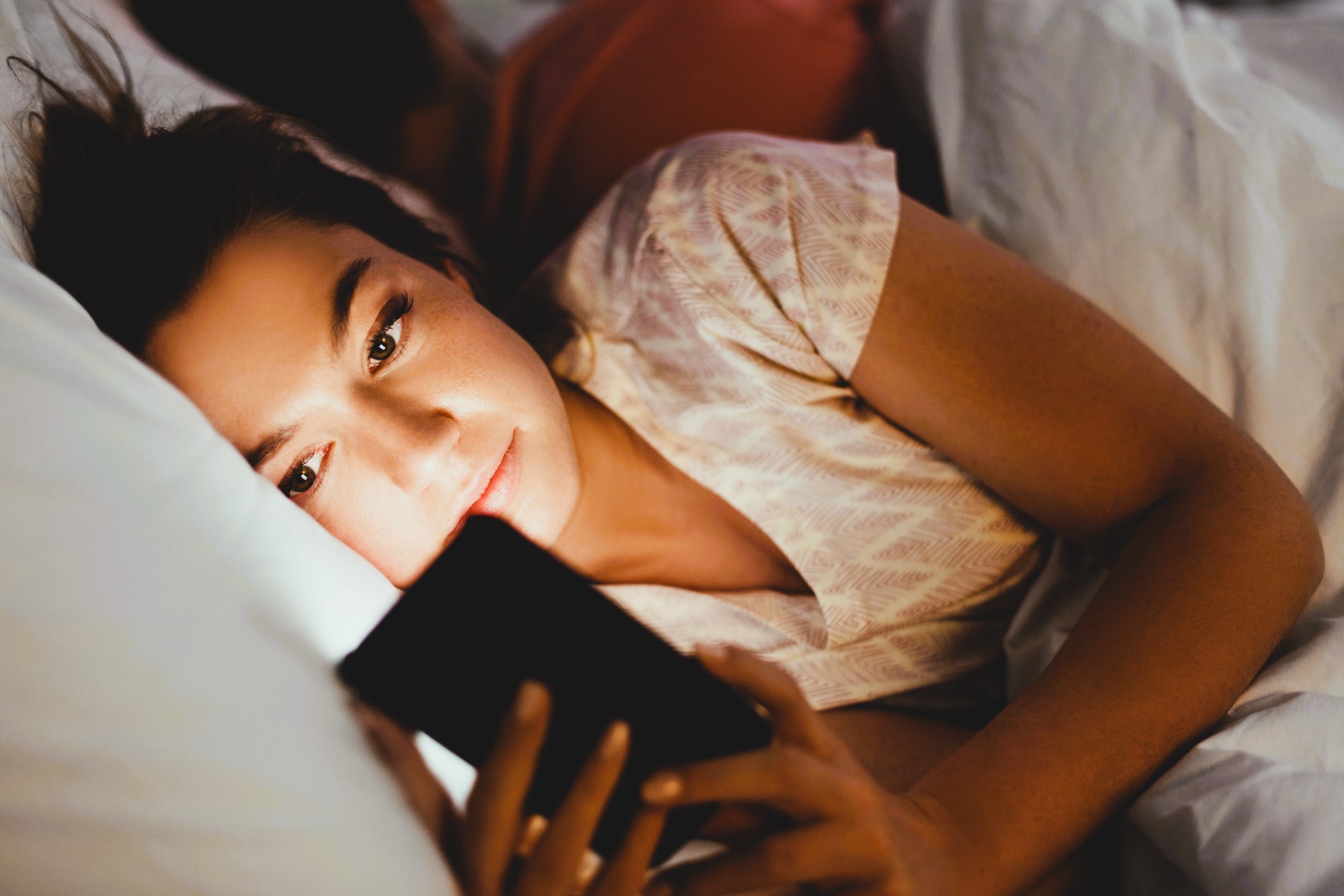 A woman is lying in bed at night, holding and looking at a smartphone. Her face is softly illuminated by the screen. She appears relaxed, with her head resting on a pillow. Another person is lying beside her.