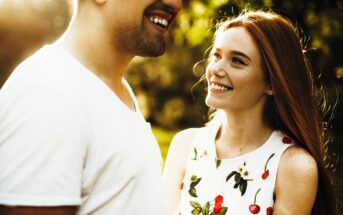 A woman with long red hair smiles at a man wearing a white T-shirt. The sunlight creates a warm glow around them. She is wearing a white dress with a floral and cherry pattern, and they are outdoors with blurred greenery in the background.