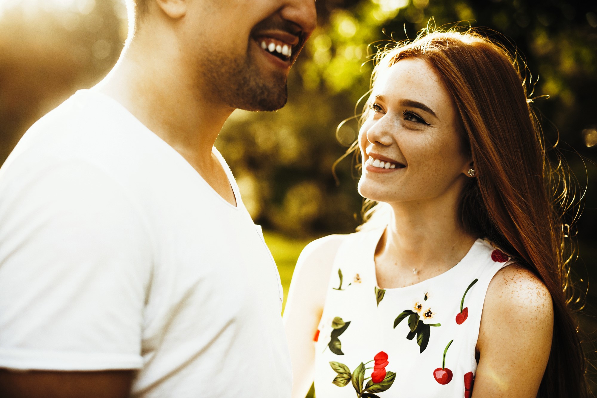 A woman with long red hair smiles at a man wearing a white T-shirt. The sunlight creates a warm glow around them. She is wearing a white dress with a floral and cherry pattern, and they are outdoors with blurred greenery in the background.