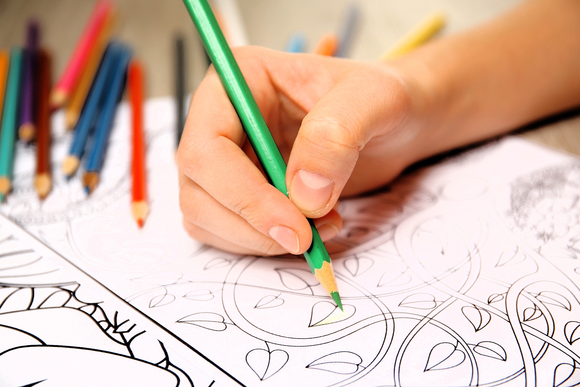 A hand coloring a leaf design with a green pencil on a black and white outline. Several colored pencils are scattered in the background.