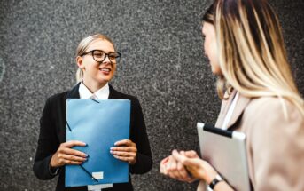 Two women engaged in conversation outside. One woman with glasses holds a blue folder, smiling. The other woman, facing away, holds a laptop. They stand against a gray textured wall.