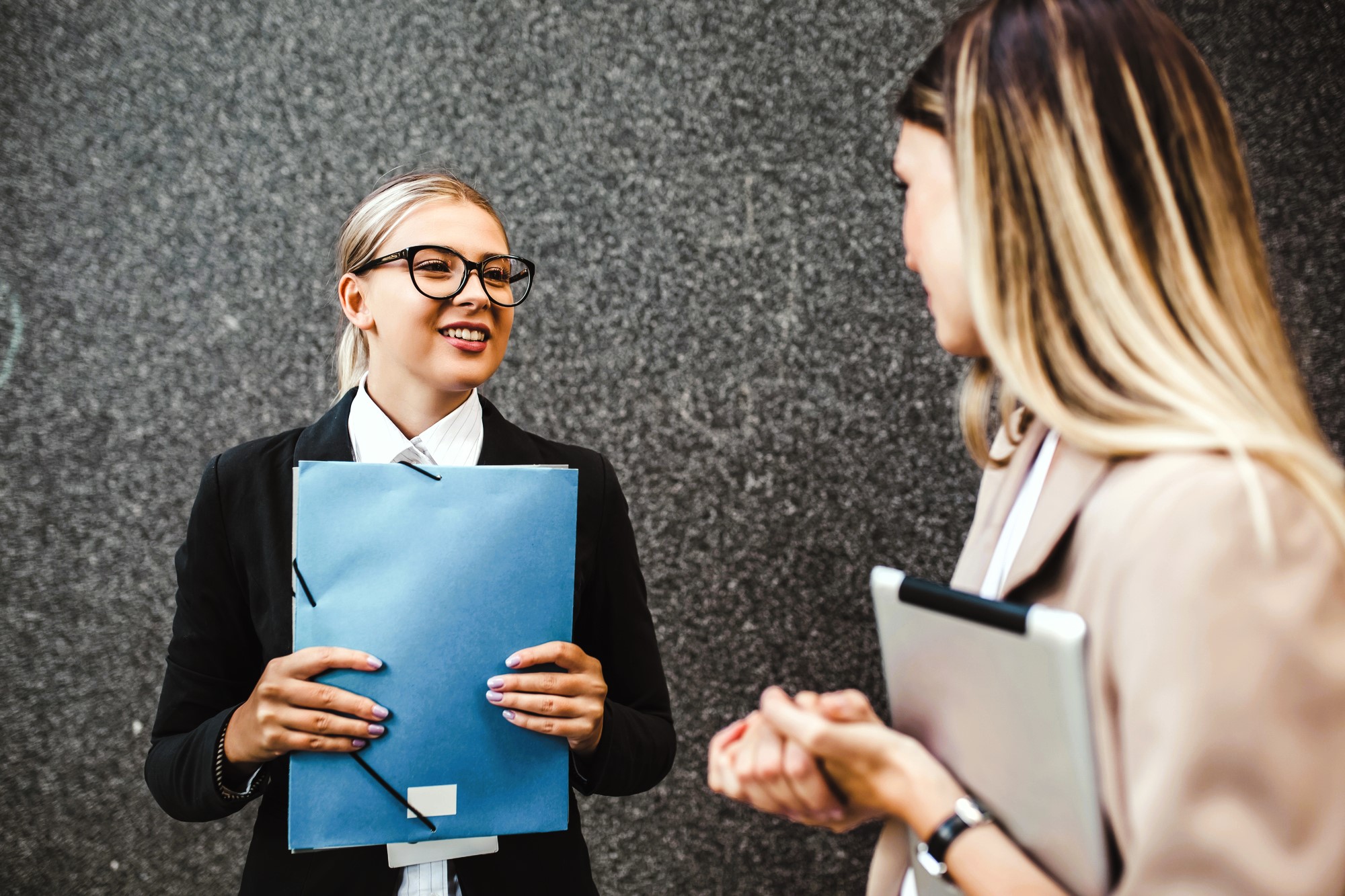 Two women engaged in conversation outside. One woman with glasses holds a blue folder, smiling. The other woman, facing away, holds a laptop. They stand against a gray textured wall.