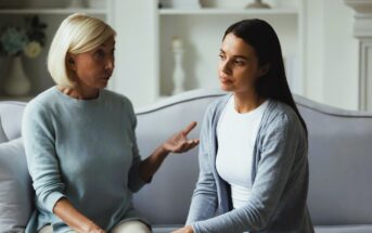 Two women sit on a sofa having a serious conversation. An older woman with short blonde hair gestures while speaking, and a younger woman with long dark hair listens intently, looking thoughtful. The background features shelves and soft lighting.