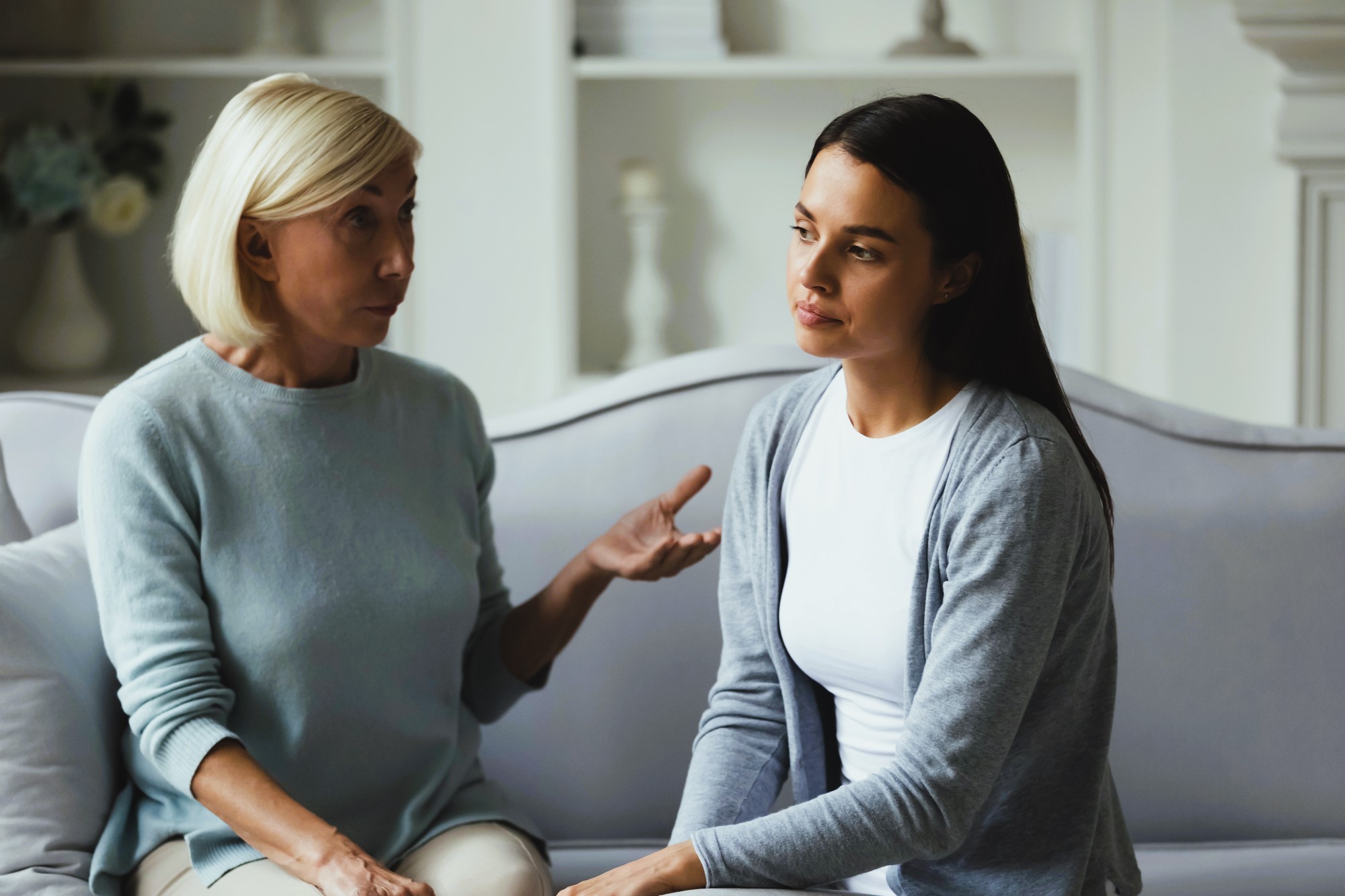 Two women sit on a sofa having a serious conversation. An older woman with short blonde hair gestures while speaking, and a younger woman with long dark hair listens intently, looking thoughtful. The background features shelves and soft lighting.