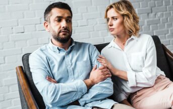 A woman with blonde hair sits on a couch next to a man with short dark hair who has his arms crossed. She gently touches his arm while looking at him, and he looks forward with a serious expression. The background is a light brick wall.