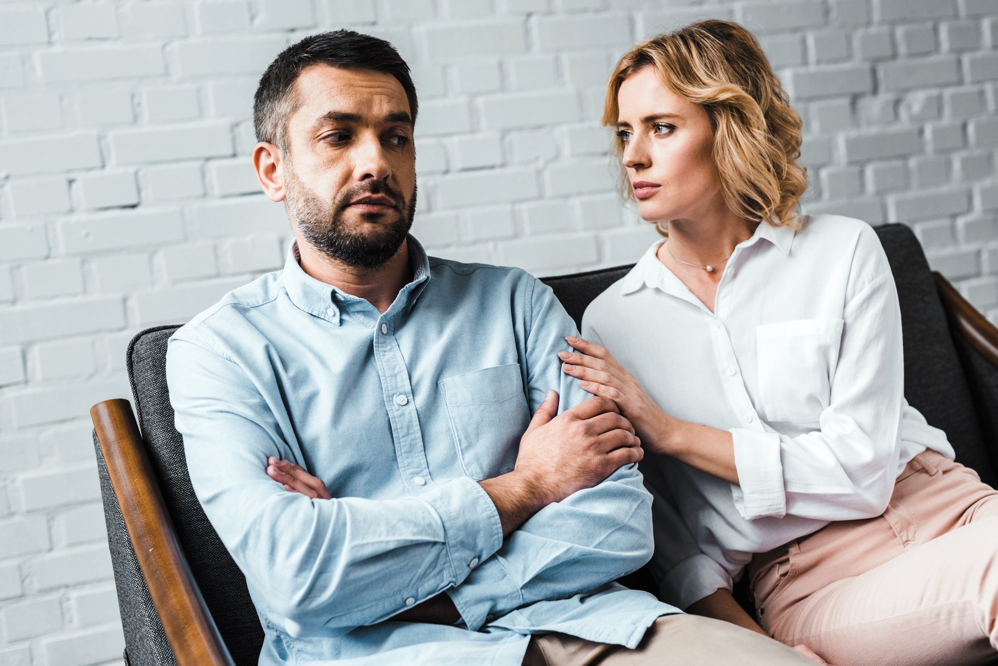 A woman with blonde hair sits on a couch next to a man with short dark hair who has his arms crossed. She gently touches his arm while looking at him, and he looks forward with a serious expression. The background is a light brick wall.