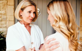 Two women with light hair stand close, looking at each other. The older woman, in a white blouse, gently holds the younger woman's arms. The younger woman has long wavy hair, and they appear to be having a heartfelt conversation.