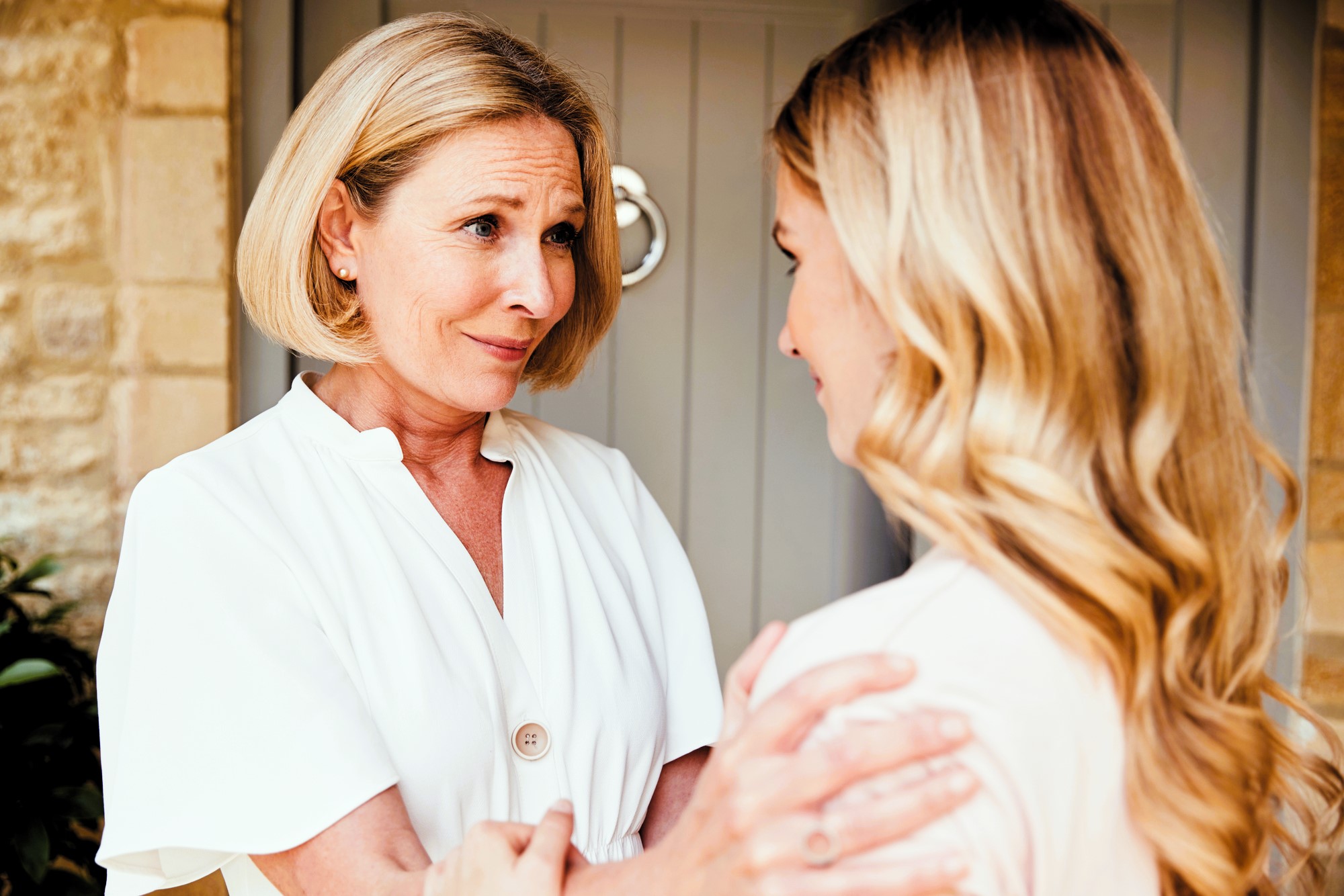 Two women with light hair stand close, looking at each other. The older woman, in a white blouse, gently holds the younger woman's arms. The younger woman has long wavy hair, and they appear to be having a heartfelt conversation.