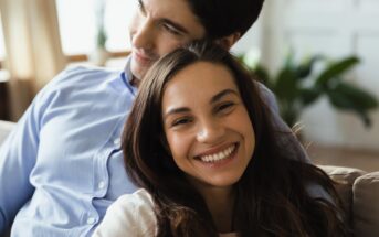 A couple sitting closely and smiling warmly indoors. The woman is in the foreground with long brown hair and a bright smile. The man is behind her, wearing a light blue shirt, looking content. The background is softly blurred with hints of greenery.