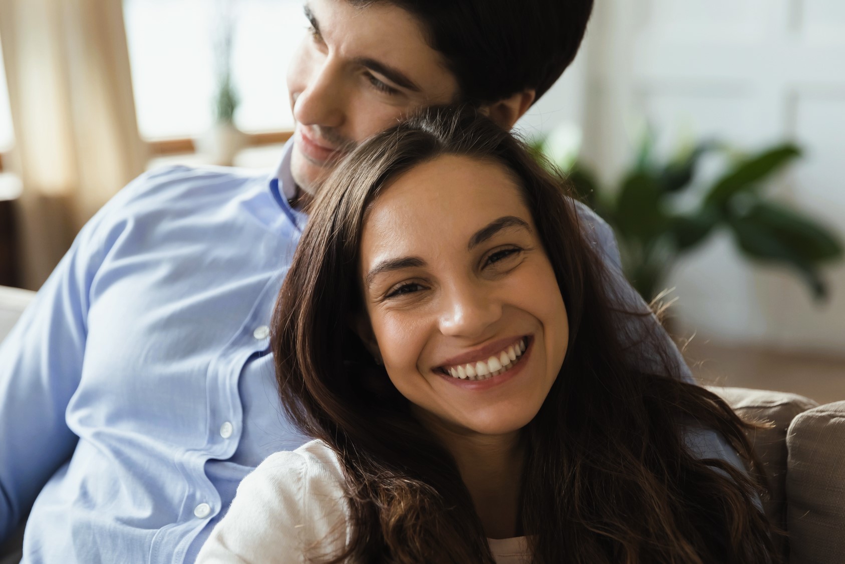 A couple sitting closely and smiling warmly indoors. The woman is in the foreground with long brown hair and a bright smile. The man is behind her, wearing a light blue shirt, looking content. The background is softly blurred with hints of greenery.