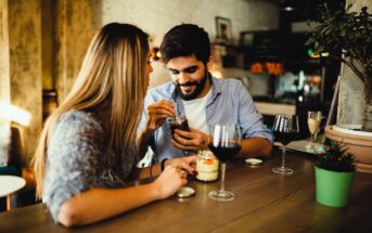 A young couple sits at a wooden table in a cozy cafe, sharing a dessert from a jar. They are smiling and enjoying the moment. Glasses of red wine and small plants decorate the table, creating a warm and inviting atmosphere.