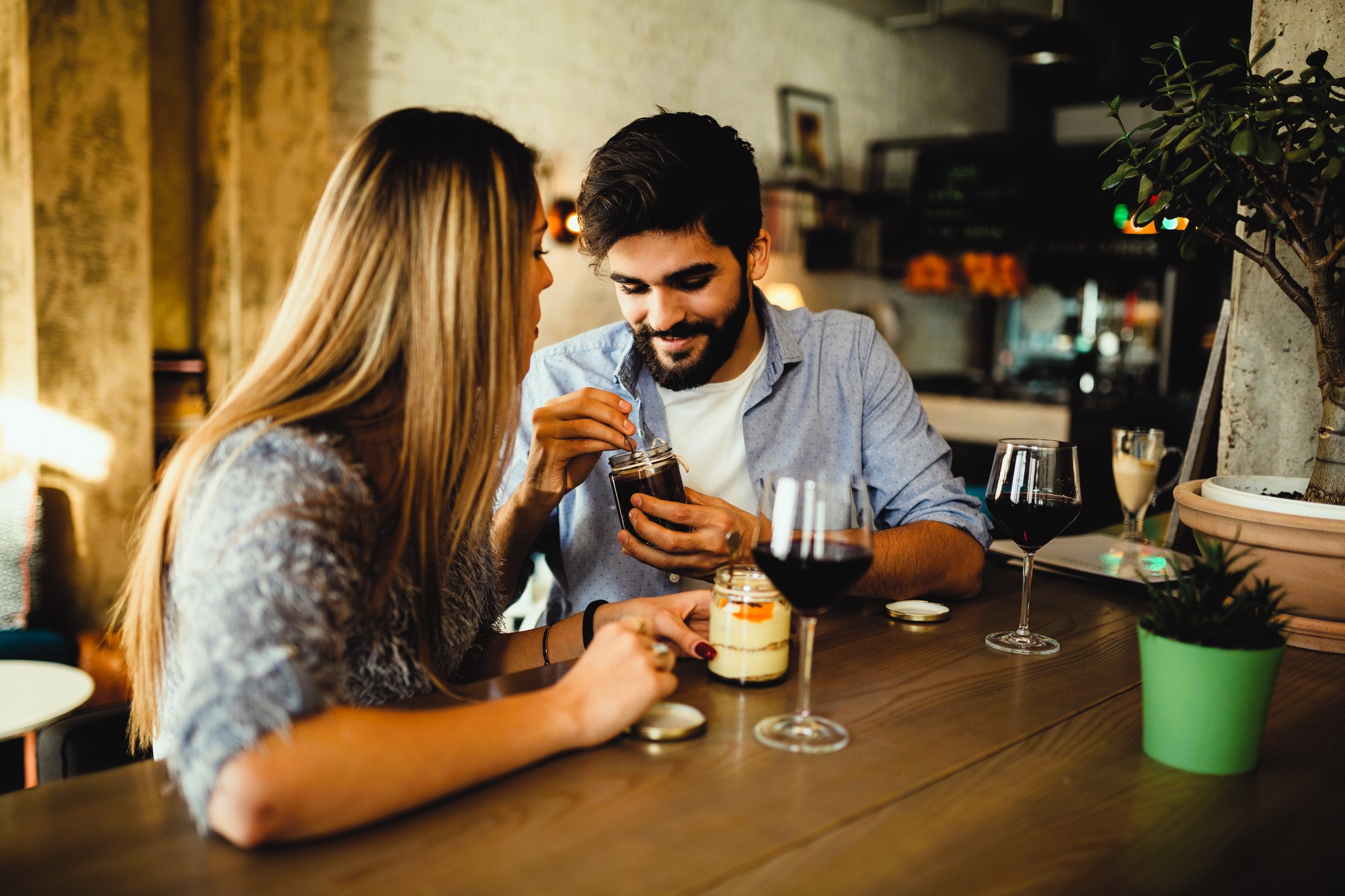 A young couple sits at a wooden table in a cozy cafe, sharing a dessert from a jar. They are smiling and enjoying the moment. Glasses of red wine and small plants decorate the table, creating a warm and inviting atmosphere.