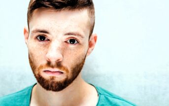 A man with short brown hair, a beard, and freckles is looking directly at the camera. He is wearing a teal shirt and is set against a light background, giving the image a bright and clear appearance.