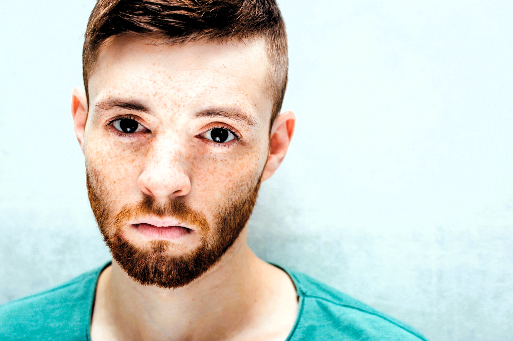 A man with short brown hair, a beard, and freckles is looking directly at the camera. He is wearing a teal shirt and is set against a light background, giving the image a bright and clear appearance.