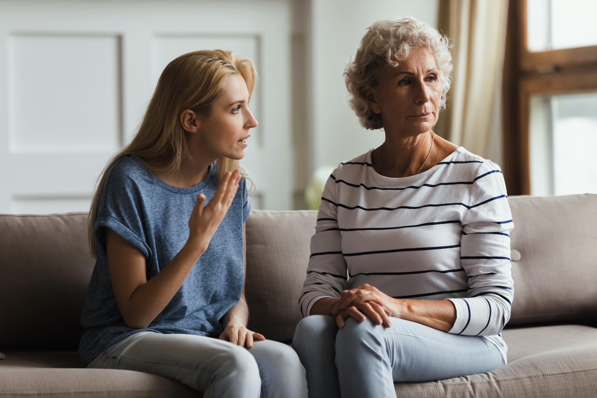 A young woman with long blonde hair talks animatedly to an older woman with curly gray hair who sits beside her, listening intently. They are sitting on a gray sofa in a bright room with a white wall and window in the background.