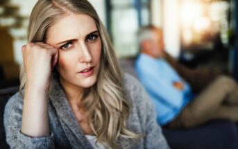 A woman with long blonde hair looks concerned, resting her head on her hand. In the background, a man with gray hair and a blue shirt sits on a couch, blurred and with arms crossed. The setting appears to be a living room.