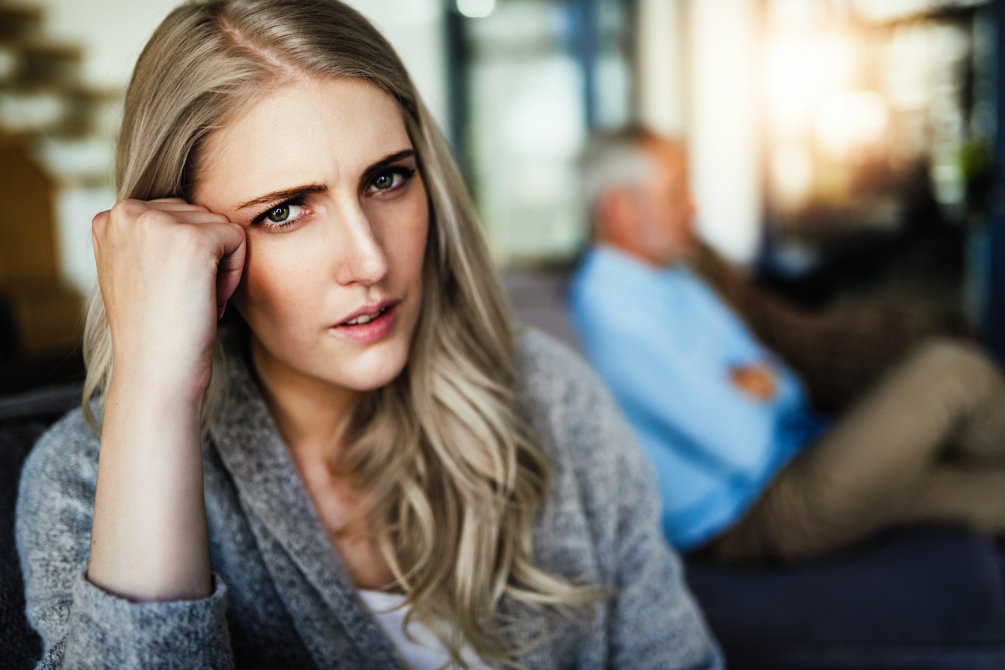 A woman with long blonde hair looks concerned, resting her head on her hand. In the background, a man with gray hair and a blue shirt sits on a couch, blurred and with arms crossed. The setting appears to be a living room.