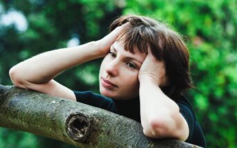 A person with short brown hair is resting their head on their hands, leaning on a tree branch with a contemplative expression. The background is blurred greenery, suggesting a natural outdoor setting.