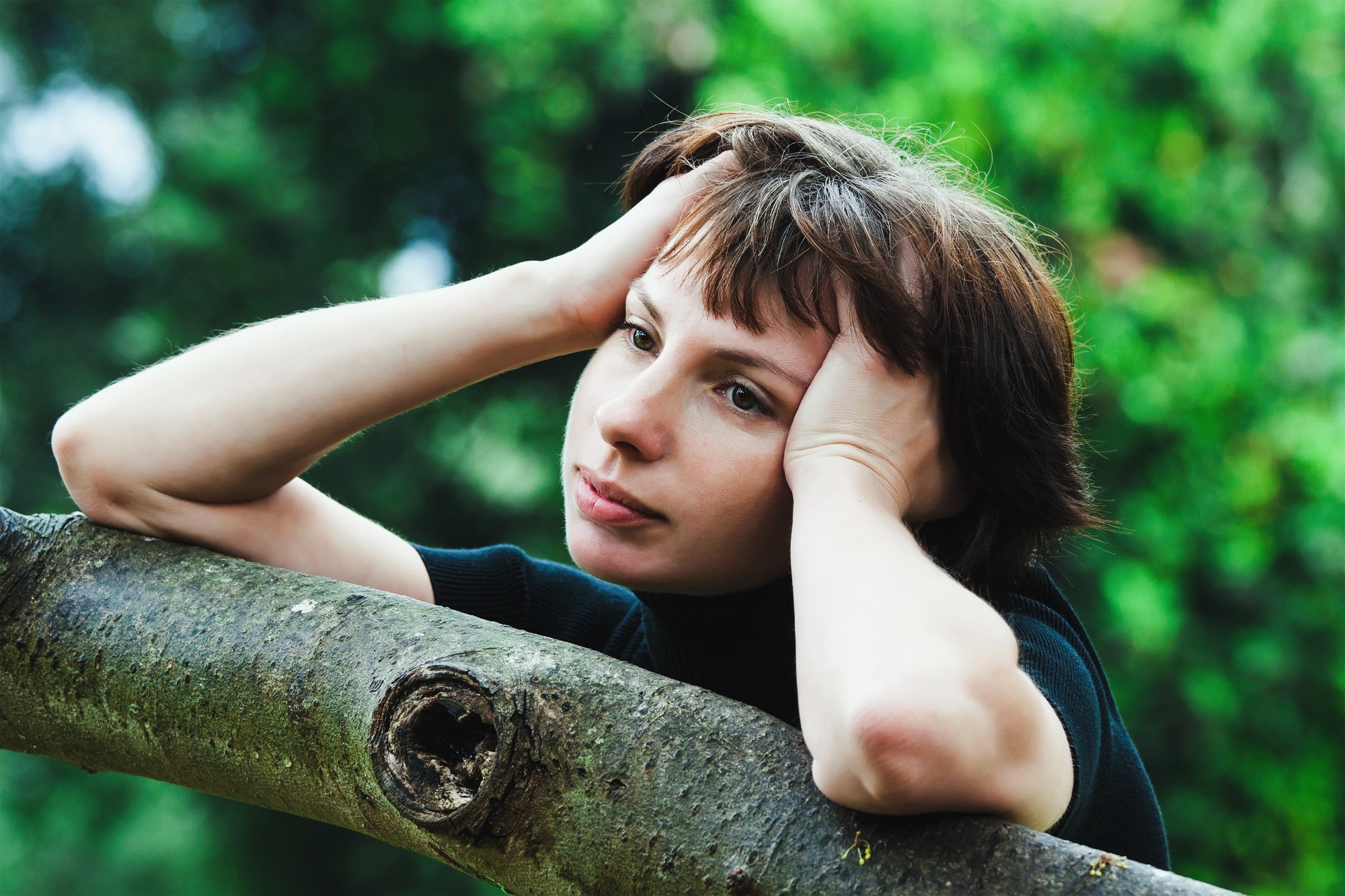 A person with short brown hair is resting their head on their hands, leaning on a tree branch with a contemplative expression. The background is blurred greenery, suggesting a natural outdoor setting.